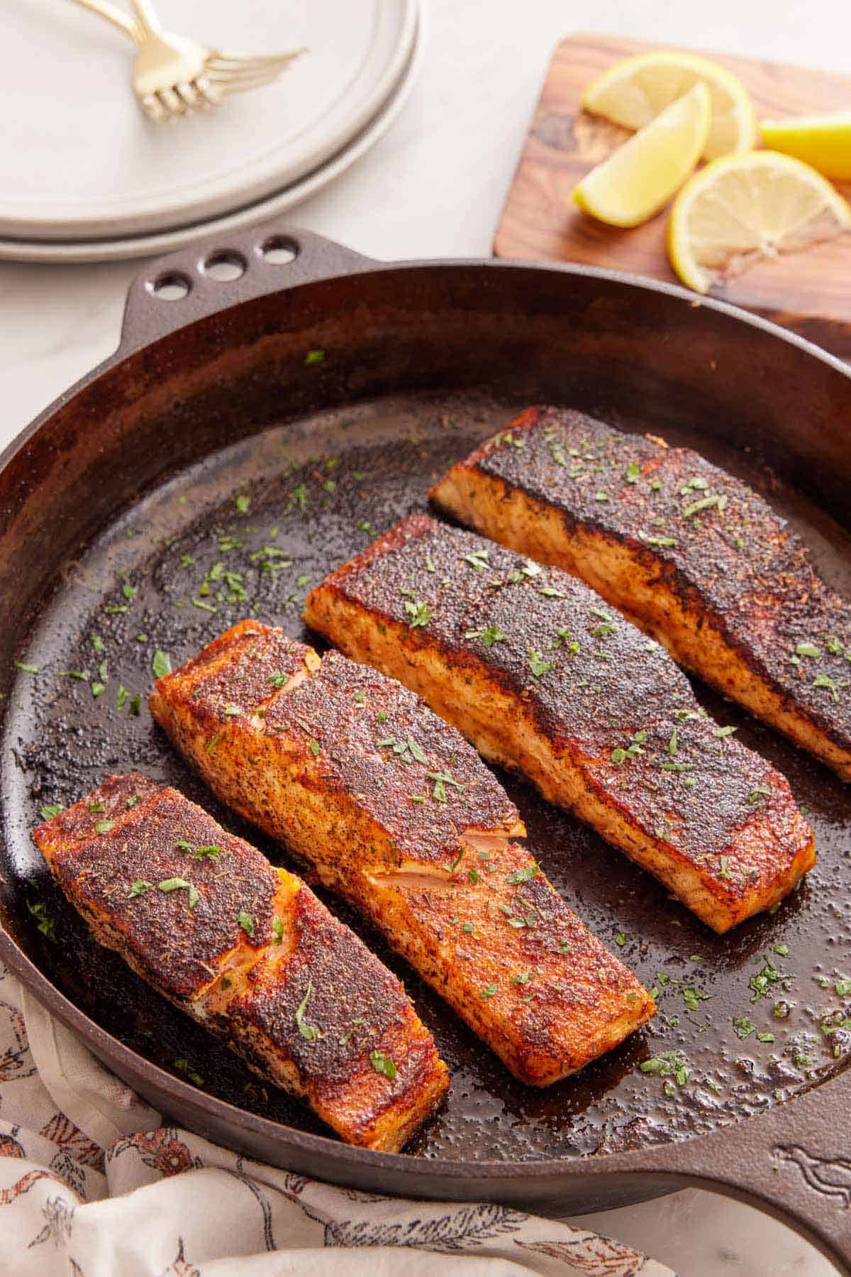 A cast iron skillet with four pieces of blackened salmon with parsley on top.