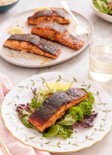 A plate with blackened salmon over salad with more salmon in the background.