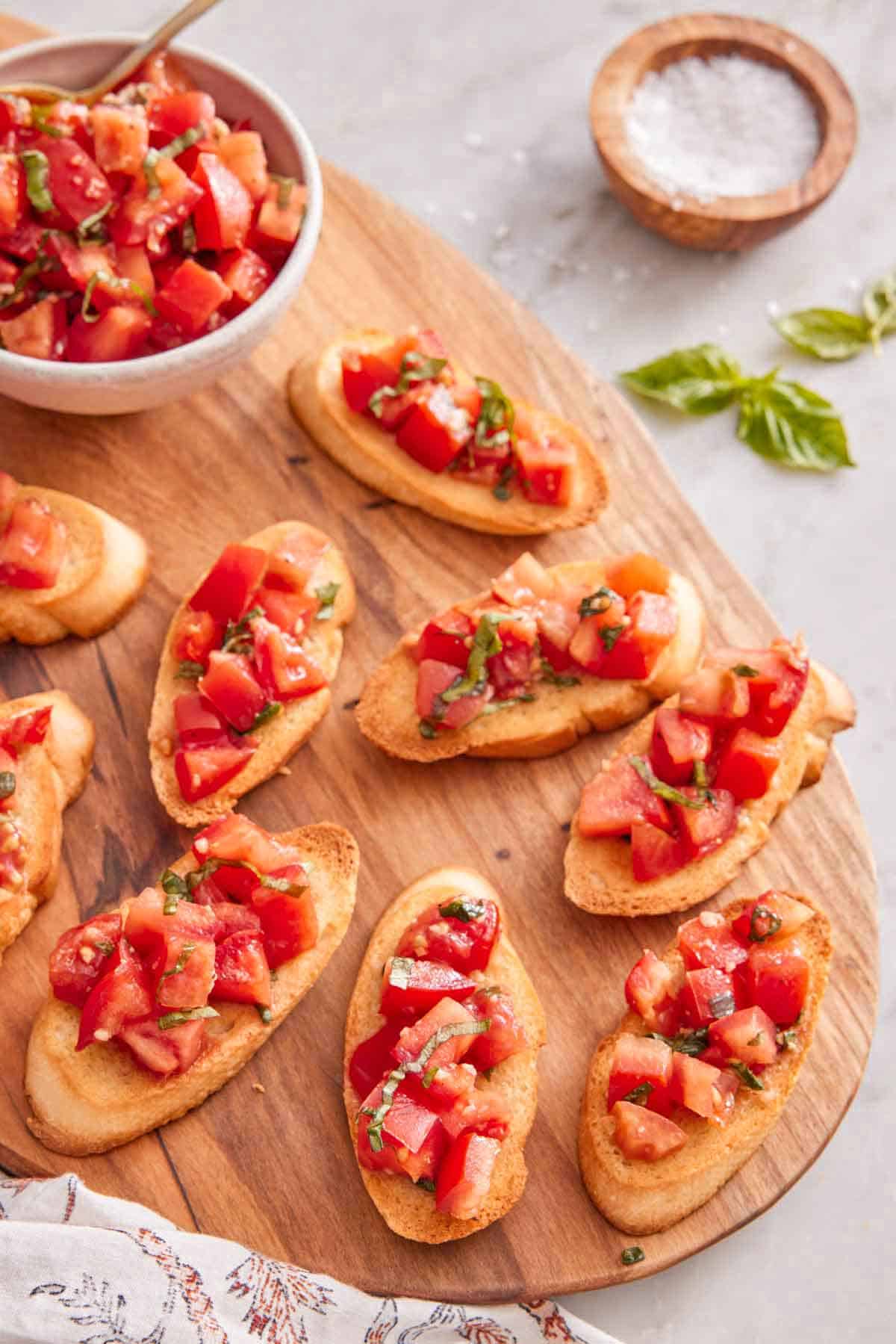 Overhead view of a wooden serving board with multiple pieces of bruschetta with a bowl of tomatoes by it.