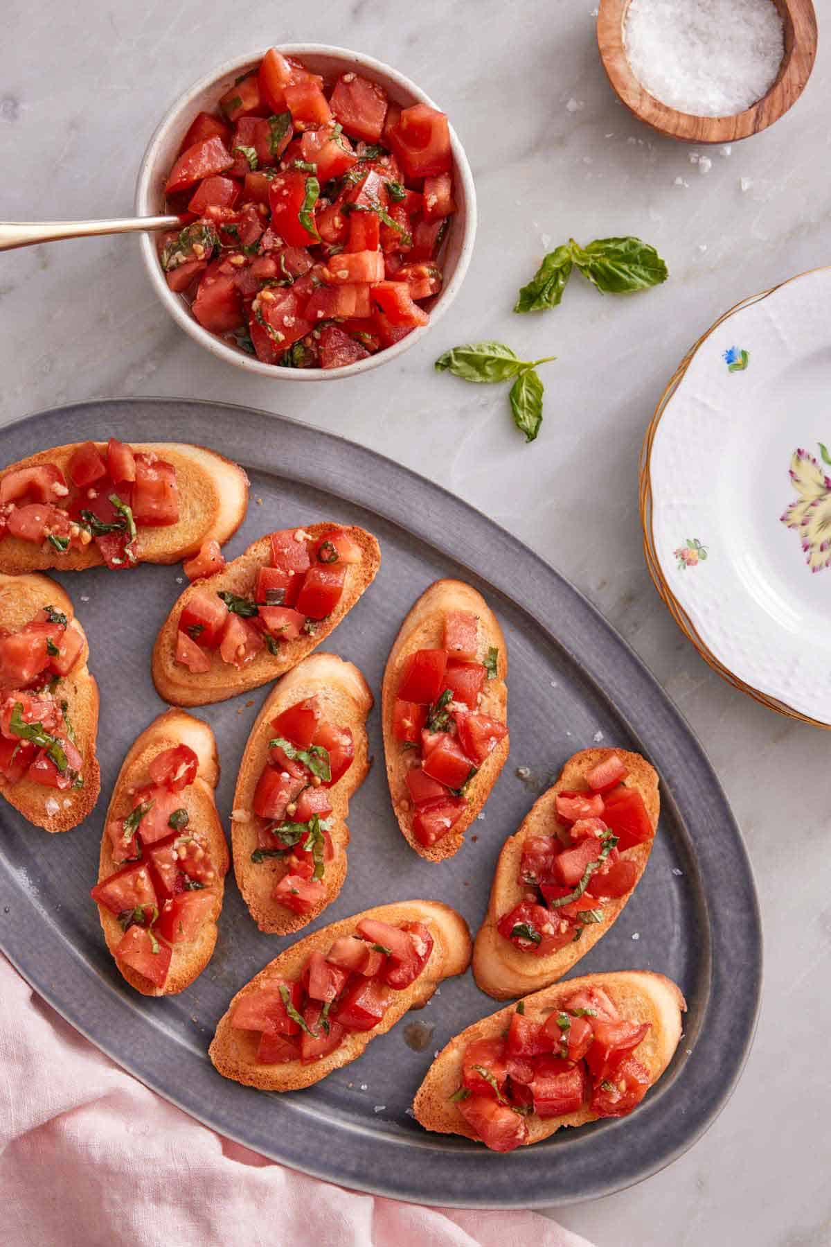 Overhead view of a grey platter with bruschetta with a bowl of tomato mixture off to the side with some salt and basil.