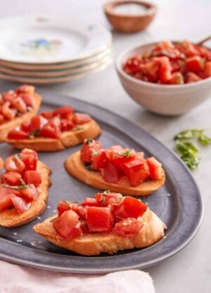 A platter with bruschetta on it with a bowl of tomato mixture in the back with a stack of plates.