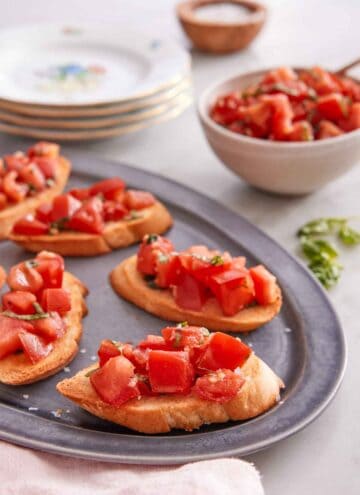 A platter with bruschetta on it with a bowl of tomato mixture in the back with a stack of plates.