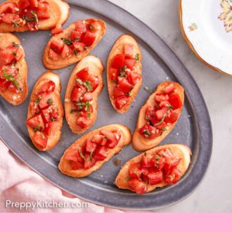 Pinterest graphic of an overhead view of a grey platter of bruschetta with a stack of plates beside it with some scattered basil.