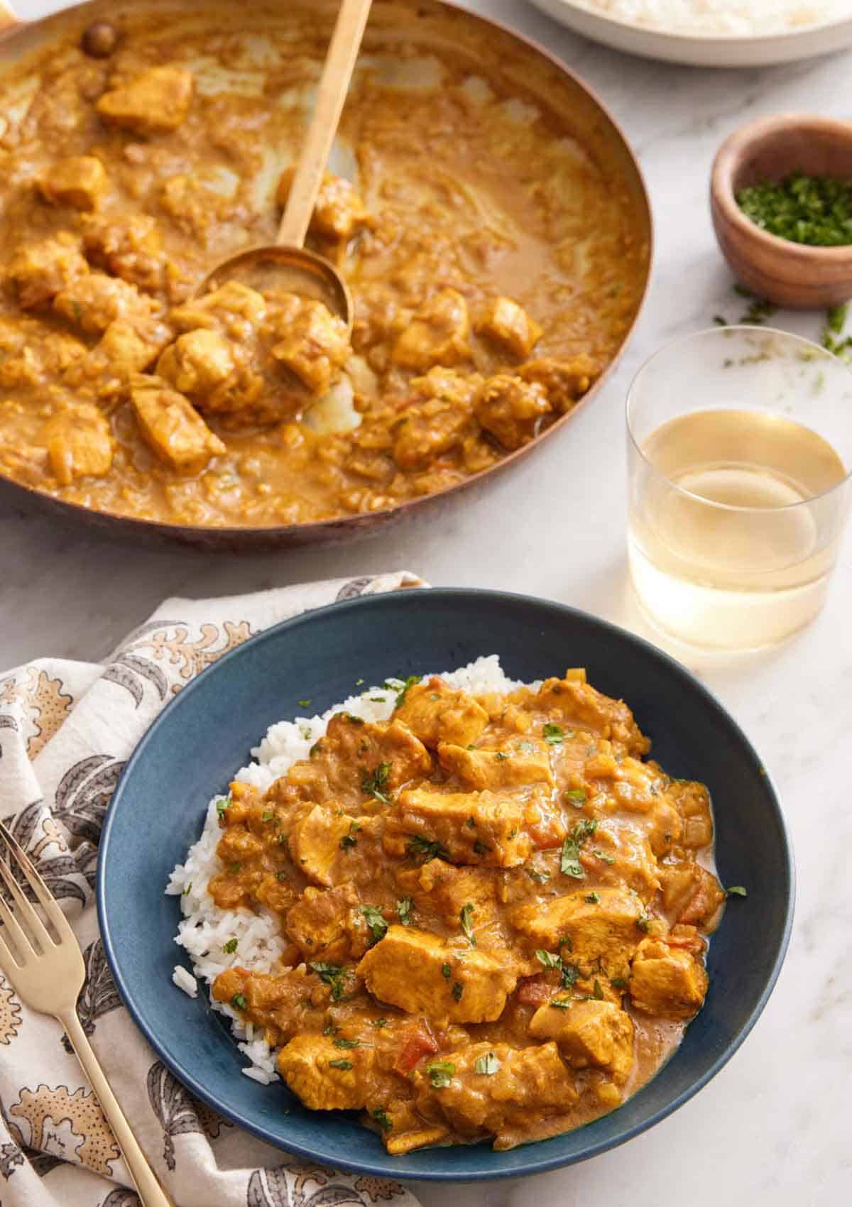 A bowl of chicken curry over rice with a skillet of more curry and glass of wine in the background.