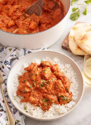 A bowl of chicken tikka masala over rice with a pot of more curry in the back along with naan.