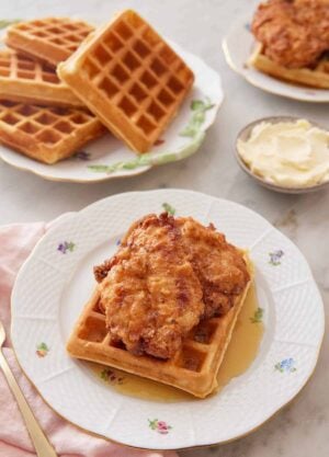 A plate with chicken and waffles with a pool of syrup with a bowl of butter and plate of waffles in the back.