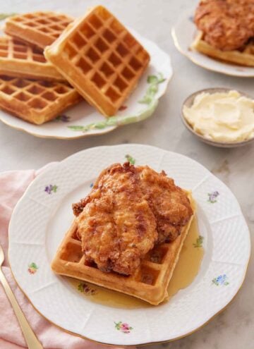 A plate with chicken and waffles with a pool of syrup with a bowl of butter and plate of waffles in the back.
