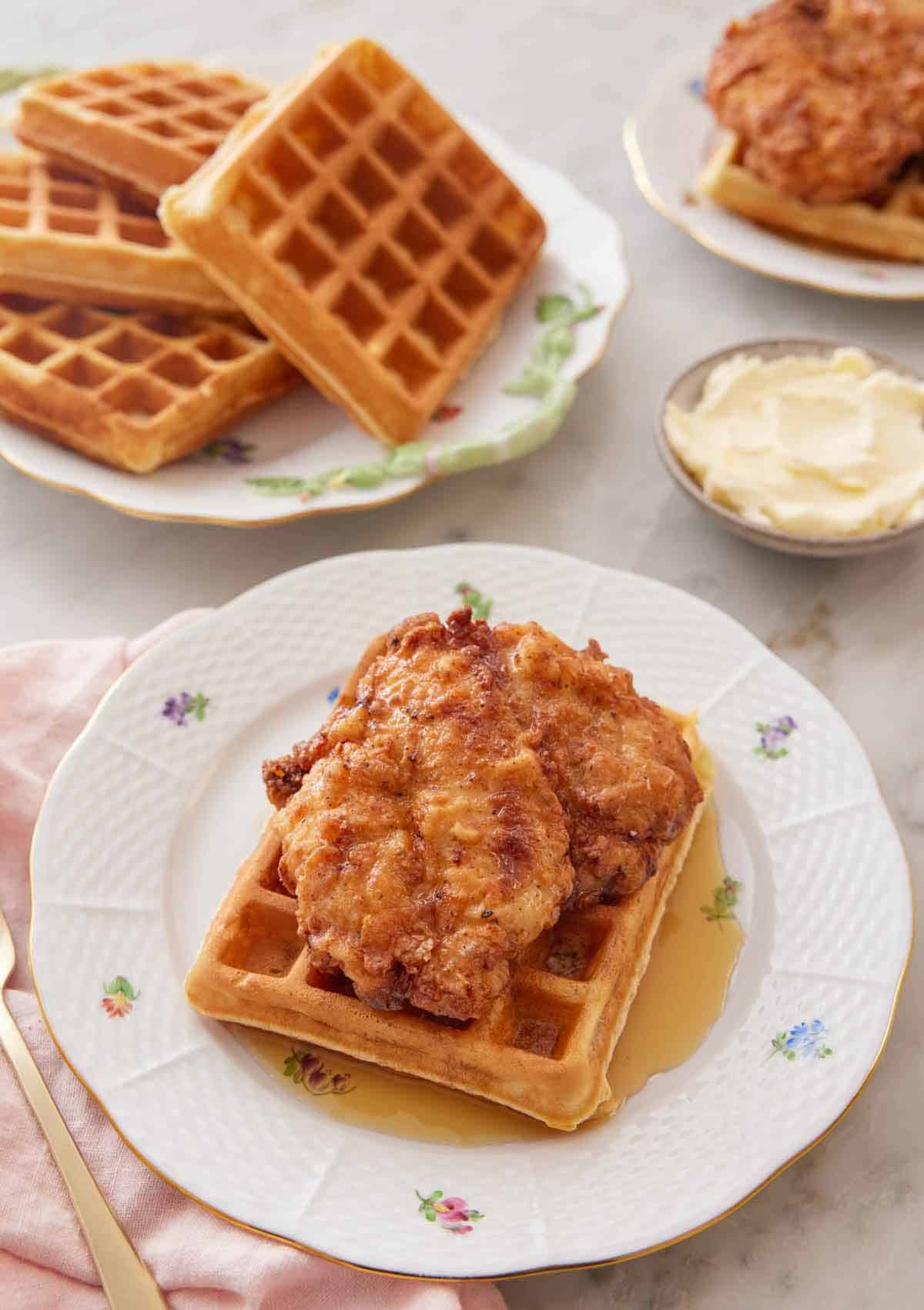 A plate with chicken and waffles with a pool of syrup with a bowl of butter and plate of waffles in the back.