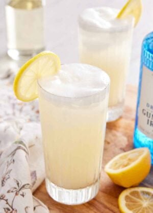 A tall glass of Gin Fizz with a lemon slice on the rim with another glass, a gin bottle, and cut lemons off to the side.