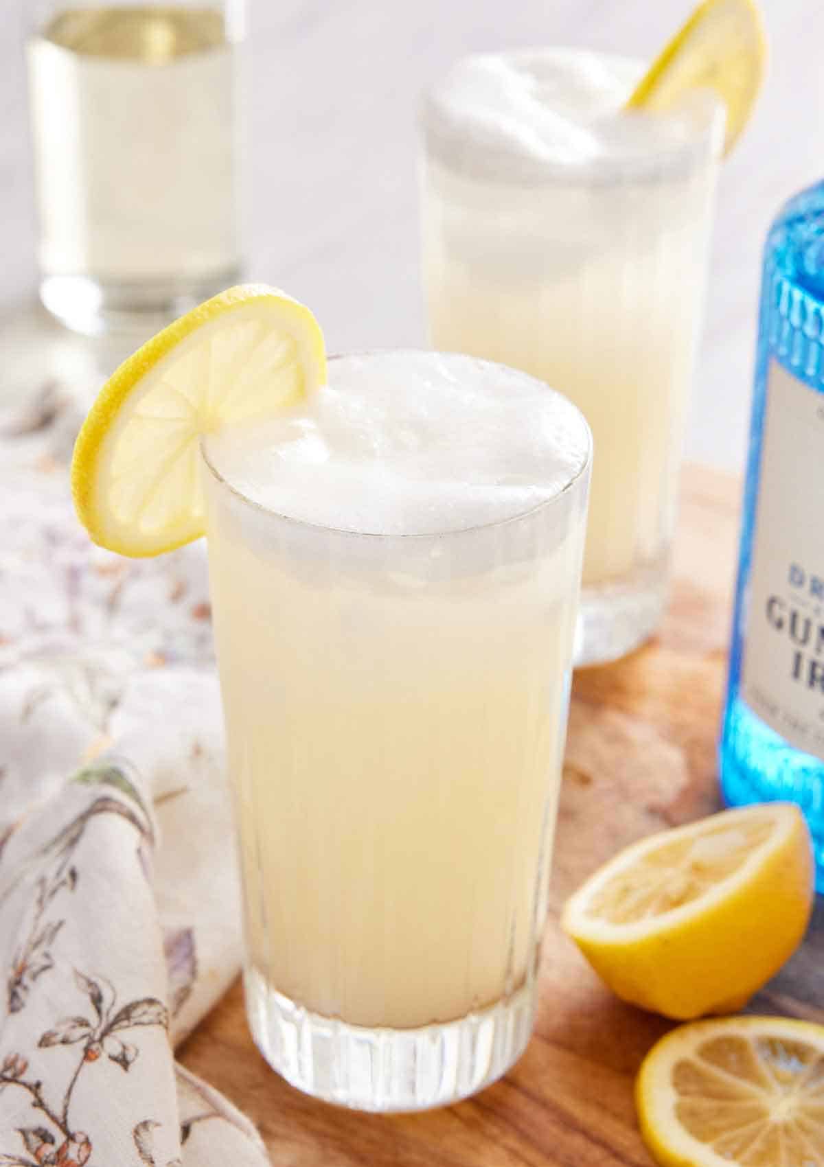 A tall glass of Gin Fizz with a lemon slice on the rim with another glass, a gin bottle, and cut lemons off to the side.
