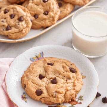 A plate with a peanut butter chocolate chip cookie with a platter of more cookies in the back along with a glass of milk.