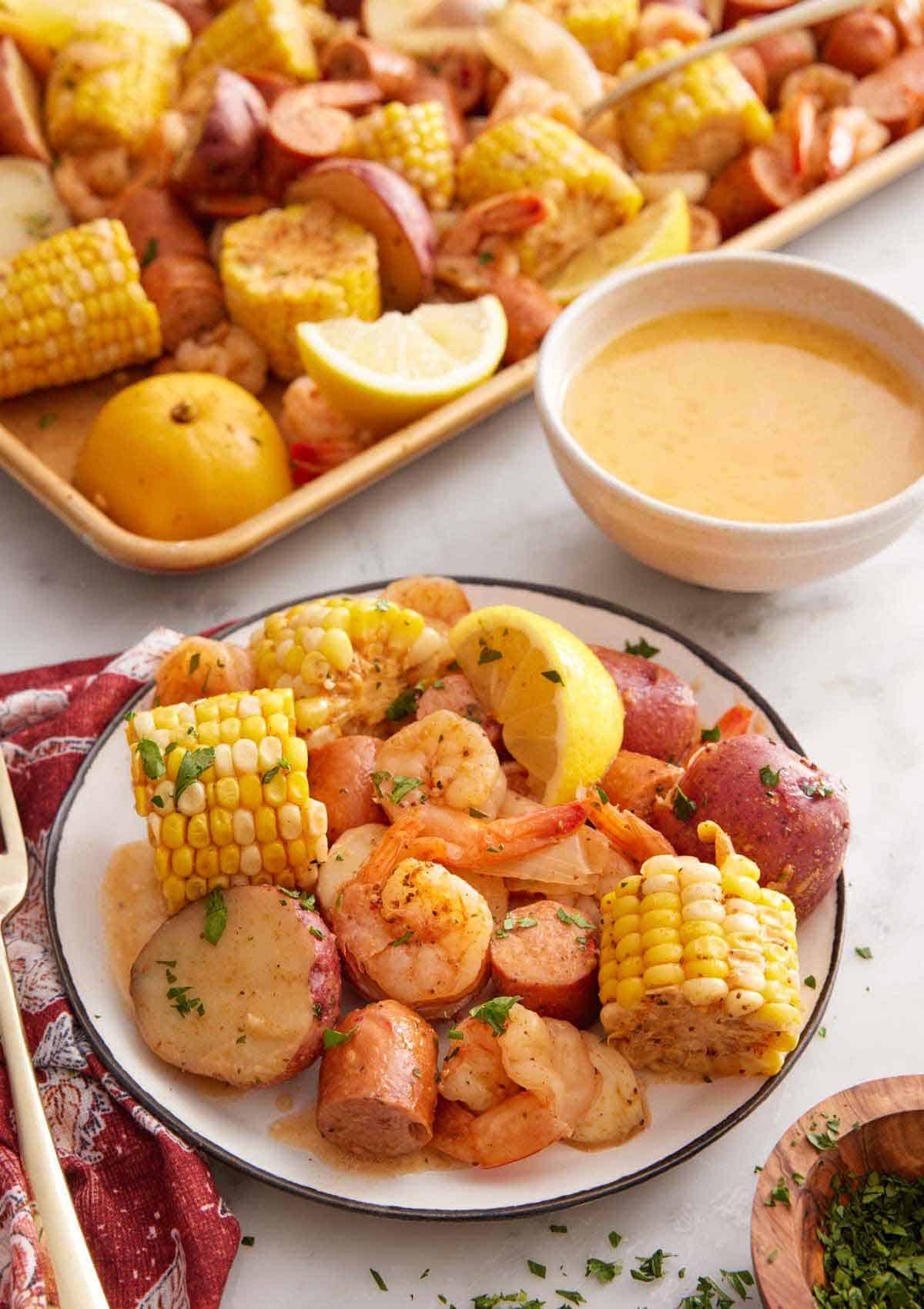 A plate of shrimp boil with a sheet pan and butter in a bowl in the back.