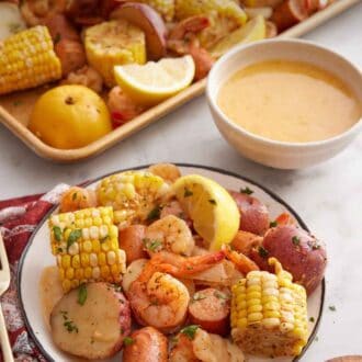 Pinterest graphic of a plate with a serving of shrimp boil with bowl of melted butter mixture and sheet pan of shrimp boil in the back.
