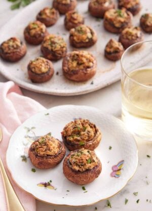 A plate with three stuffed mushrooms by a glass of wine and a platter with more mushrooms.