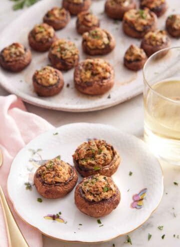 A plate with three stuffed mushrooms by a glass of wine and a platter with more mushrooms.