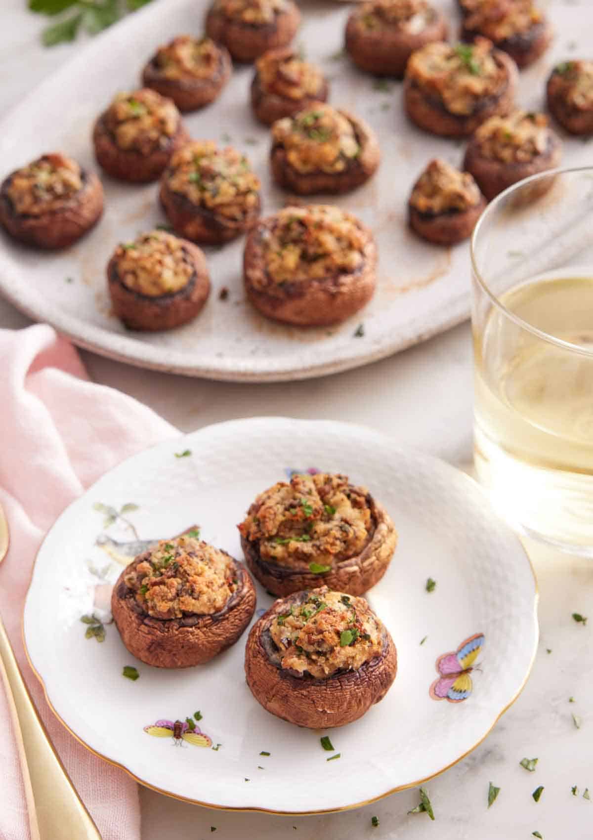 A plate with three stuffed mushrooms by a glass of wine and a platter with more mushrooms.