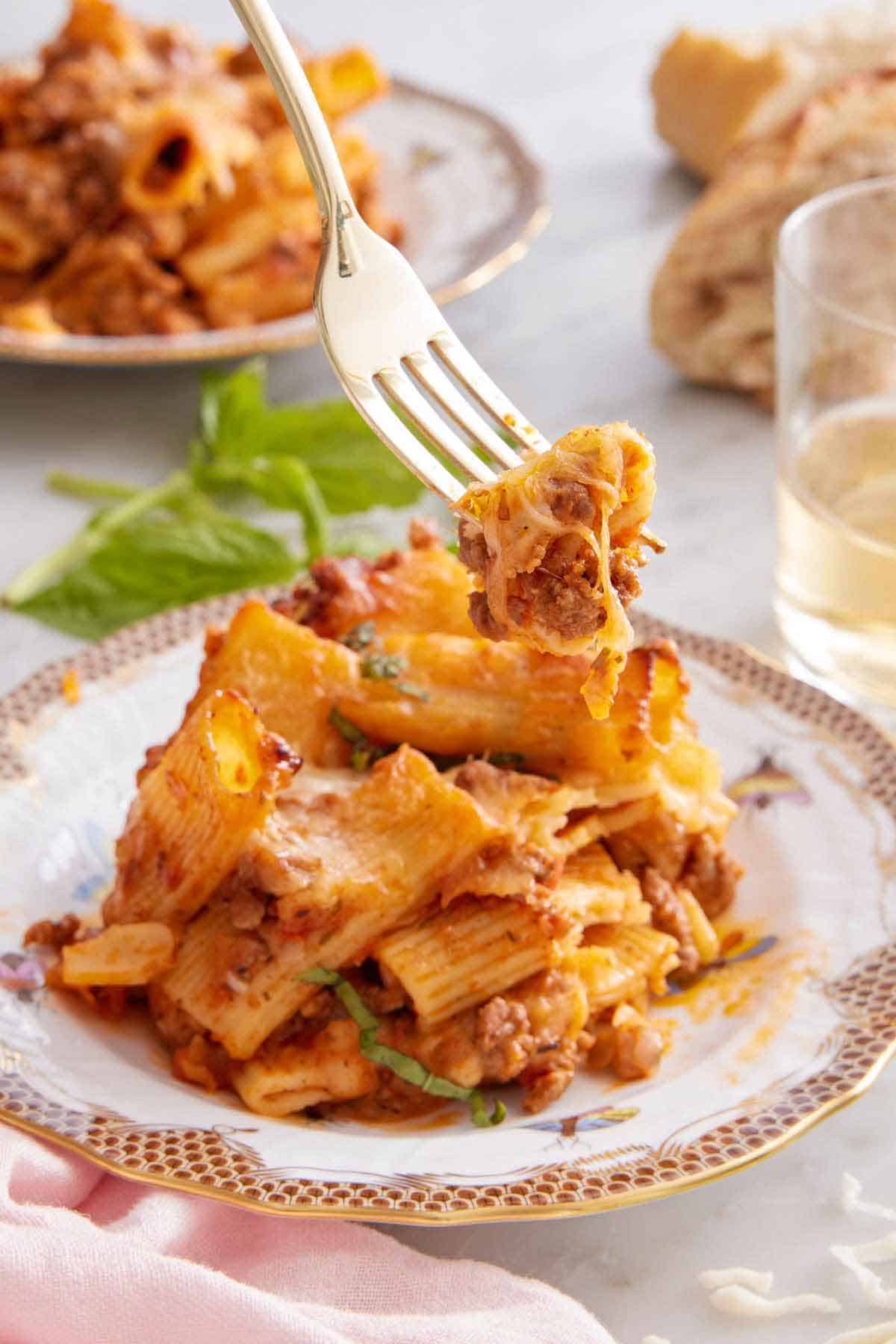 A fork lifting up a bite of baked rigatoni from a plate. A second plate, basil, wine, and bread in the background.