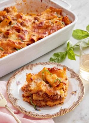 A plate with a serving of baked rigatoni with the baking dish in the back and some basil and wine to the side.
