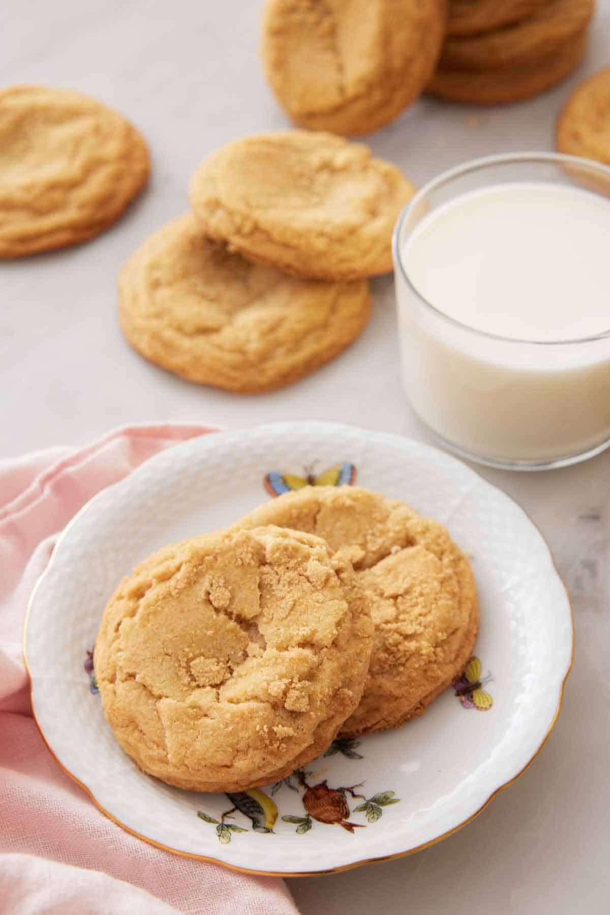 A plate with two brown sugar cookies with a glass of milk and more cookies in the back.