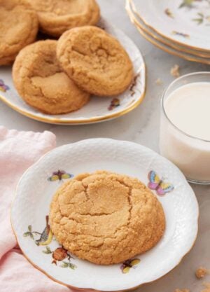 A plate with a brown sugar cookie with a platter with more cookies and a glass of milk behind it.