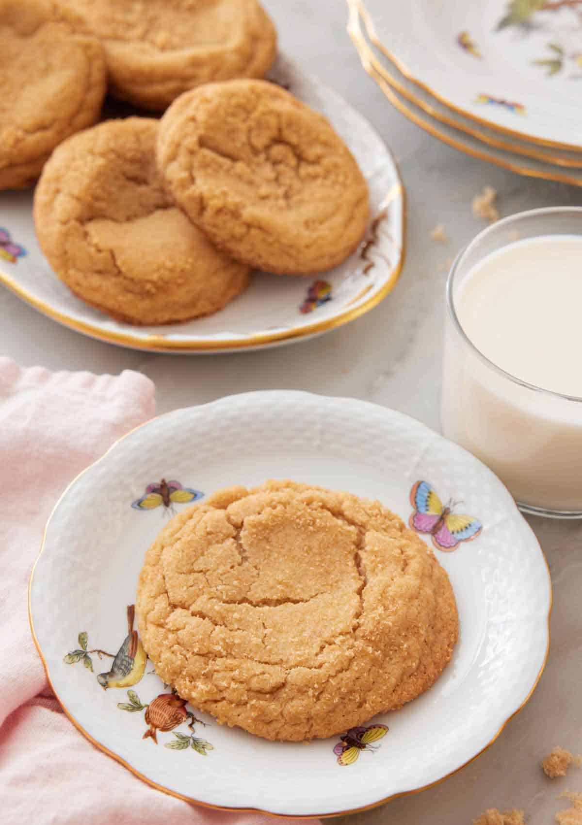 A plate with a brown sugar cookie with a platter with more cookies and a glass of milk behind it.