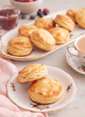 A plate with two buttermilk biscuits with a platter with more in the background along with a cup of coffee.