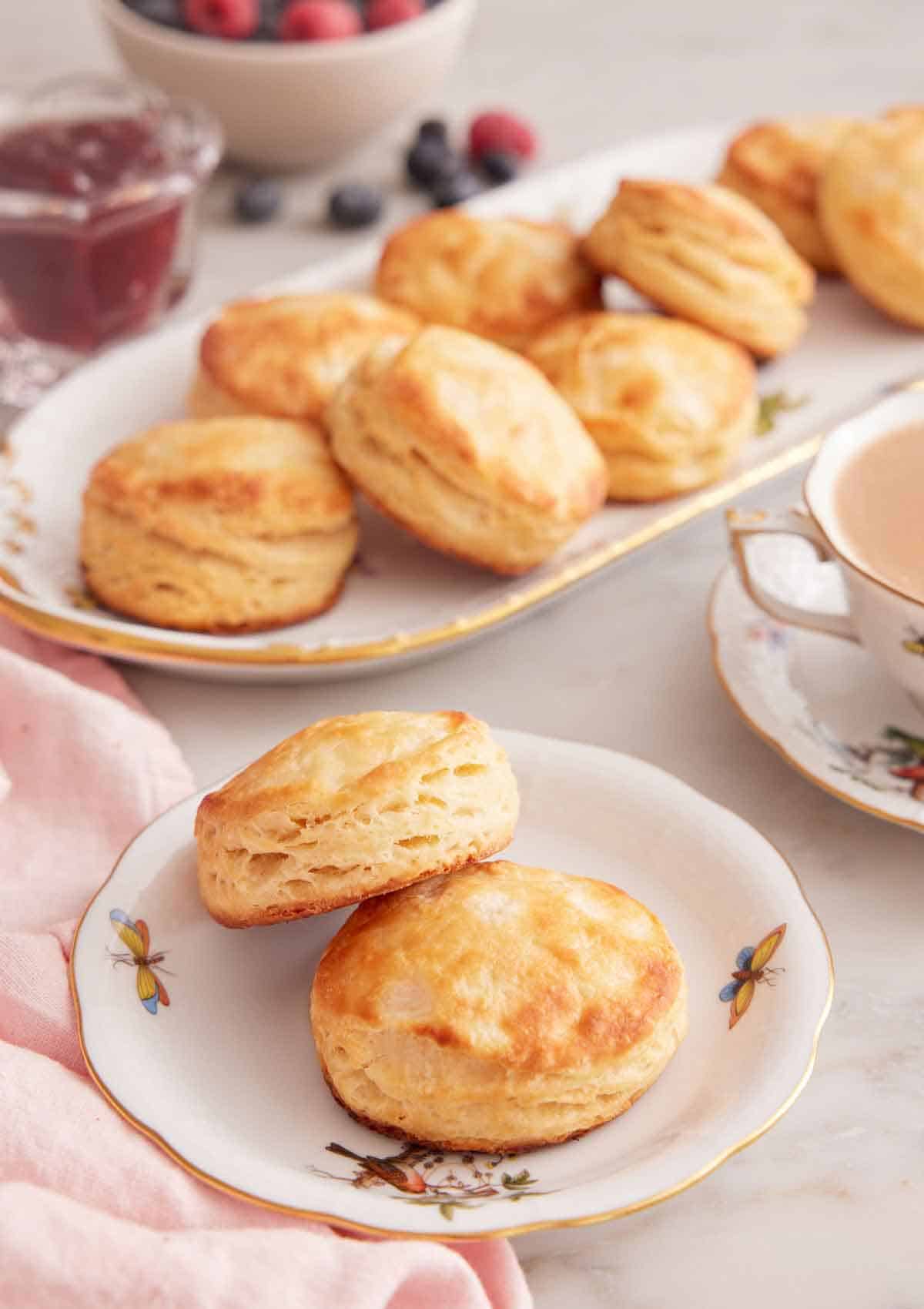A plate with two buttermilk biscuits with a platter with more in the background along with a cup of coffee.