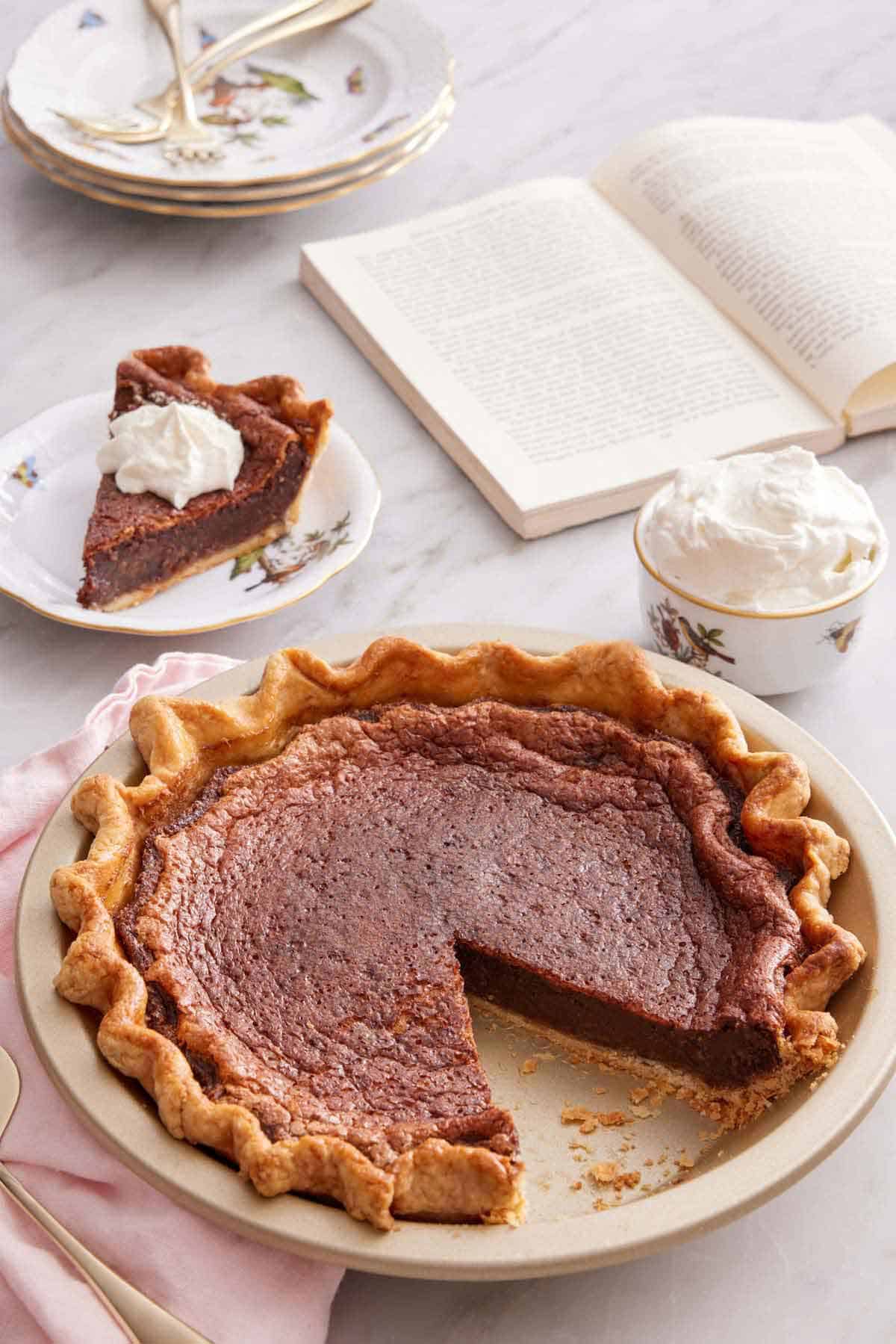 A pie dish with a chocolate chess pie with a slice cut out and plated in the background along with a bowl of whipped cream and an opened book.