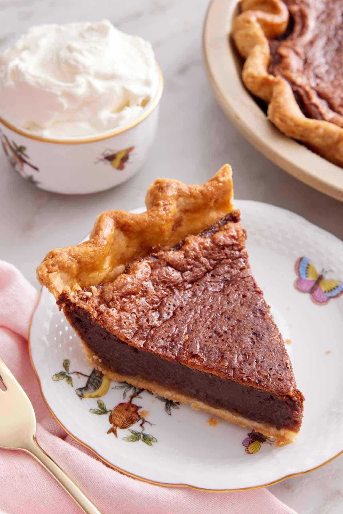A slice of chocolate chess pie on a plate with a bowl of whipped cream in the back.