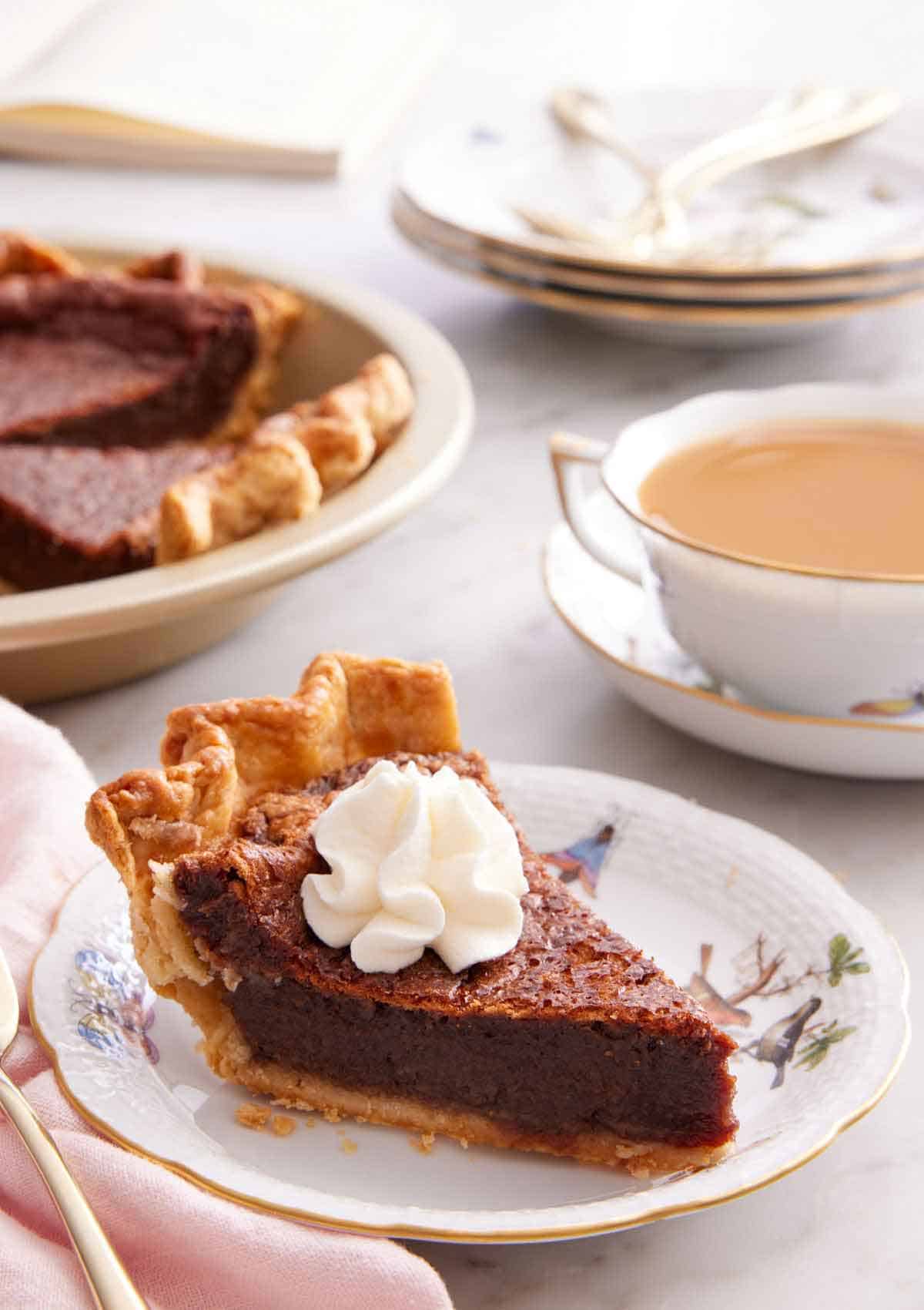 A slice of chocolate chess pie on a plate with a dollop of whipped cream on top. Coffee and more pie in the background.