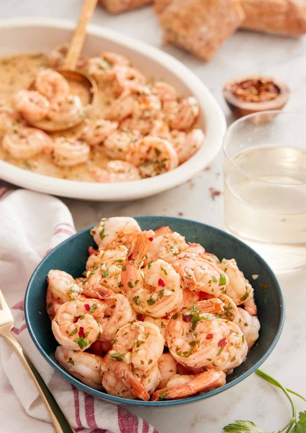 A bowl of garlic shrimp with a platter of shrimp in the back along with some wine.