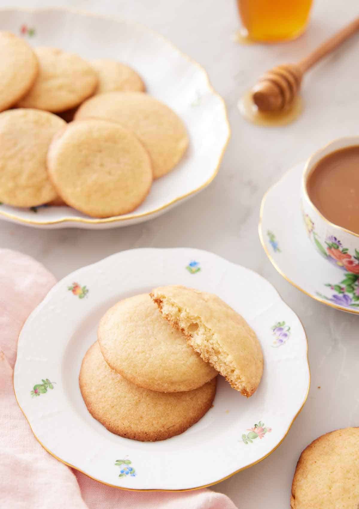 A plate with two and a half honey cookies with a platter of more cookies and cup of coffee in the back.