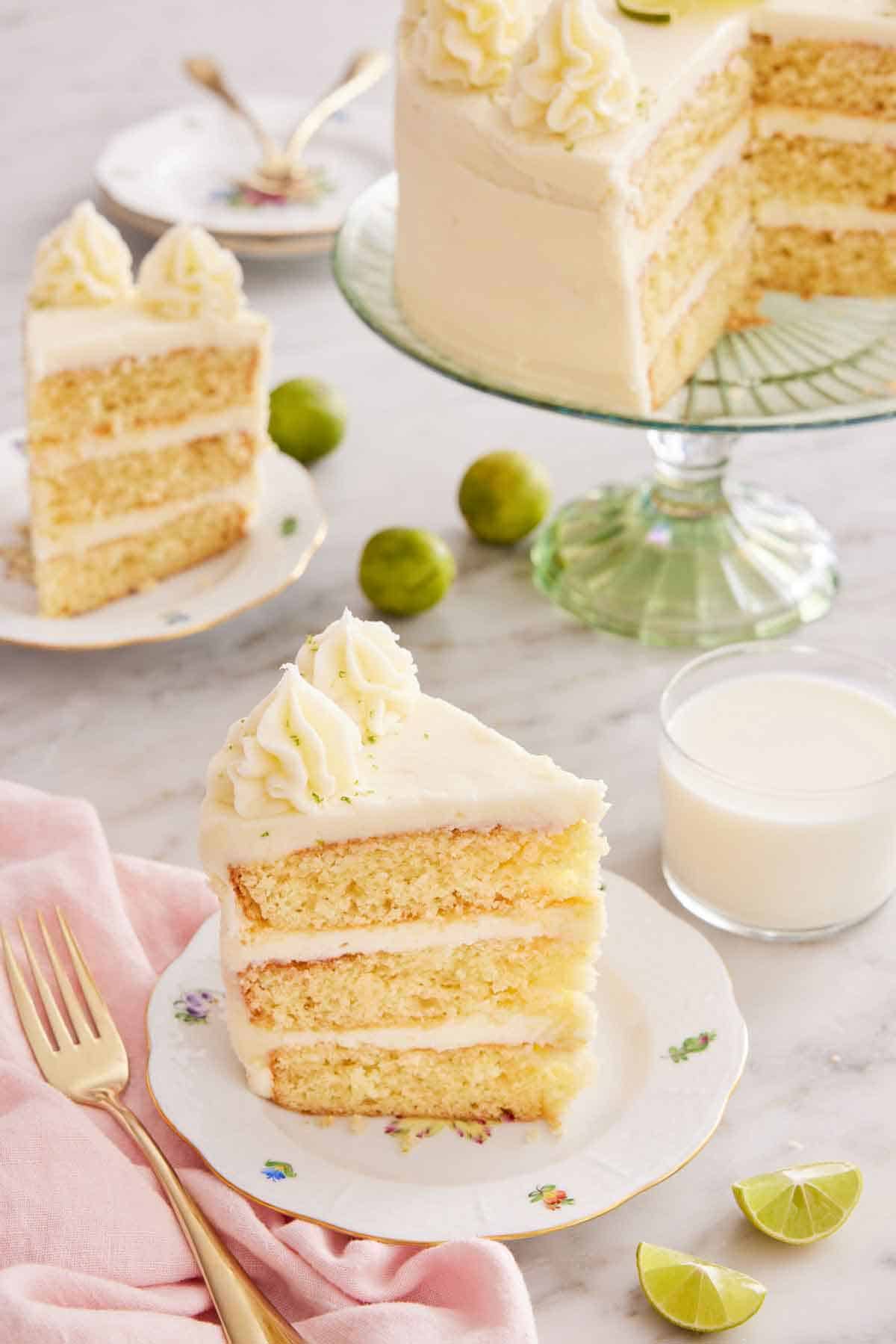 Two slices of key lime cake, plated, with a cake stand with the rest of the cut cake, a glass of milk, and cut limes in the background.