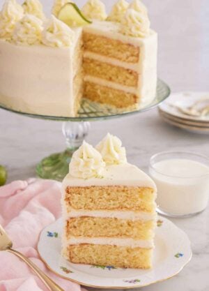 A slice of key lime cake on a plate with a glass of milk and the rest of the cake in the background.