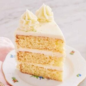 A slice of key lime cake with two dollops of frosting and lime zest on top. Glass of milk, fork, and cut limes beside it.