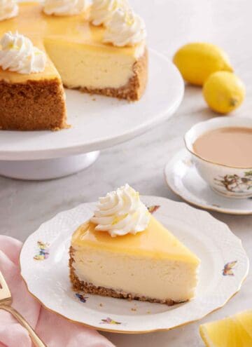 A slice of lemon cheesecake with whipped cream with a cup of coffee and the rest of the cake on a cake stand in the back.