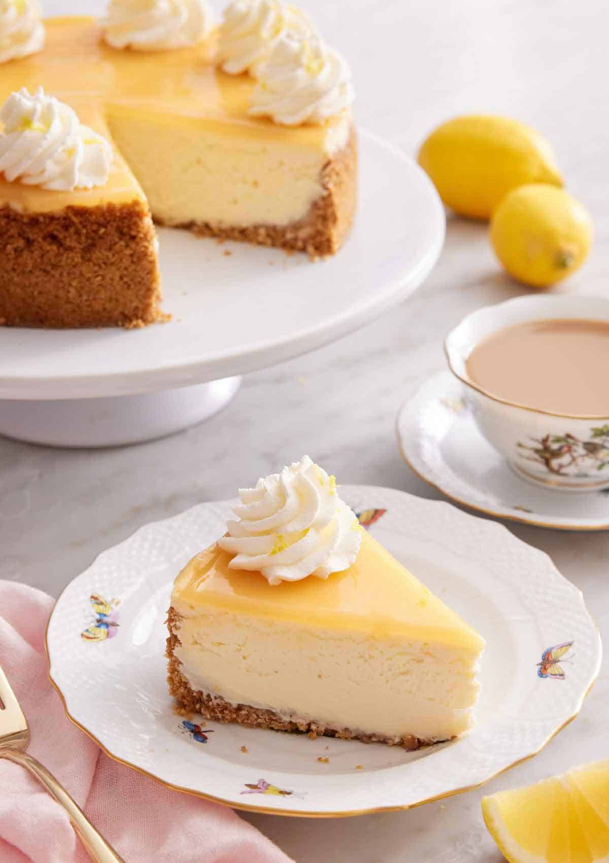 A slice of lemon cheesecake with whipped cream with a cup of coffee and the rest of the cake on a cake stand in the back.