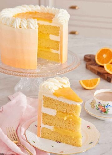 A slice of orange creamsicle cake with the rest of the cake on a cake stand in the background along with some cut oranges and a cup of coffee.