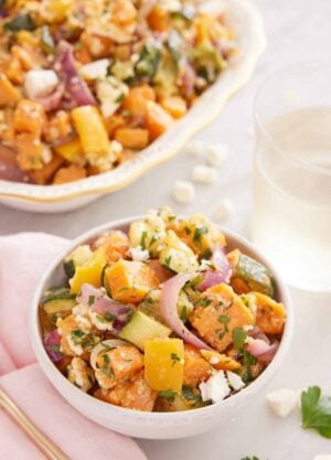 A bowl of roasted vegetable salad with a glass of wine and platter in the back.
