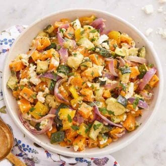 A large bowl of roasted vegetable salad with some crumbled feta, a linen napkin and large spoon on the side.