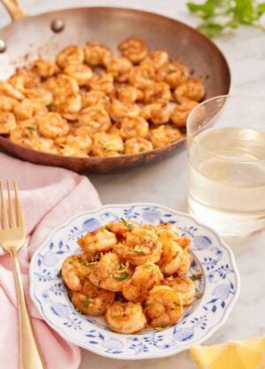 A plate of sautéed shrimp with a glass of wine and skillet of shrimp in the back.