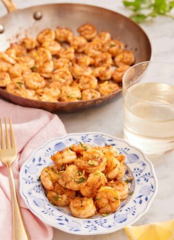 A plate of sautéed shrimp with a glass of wine and skillet of shrimp in the back.