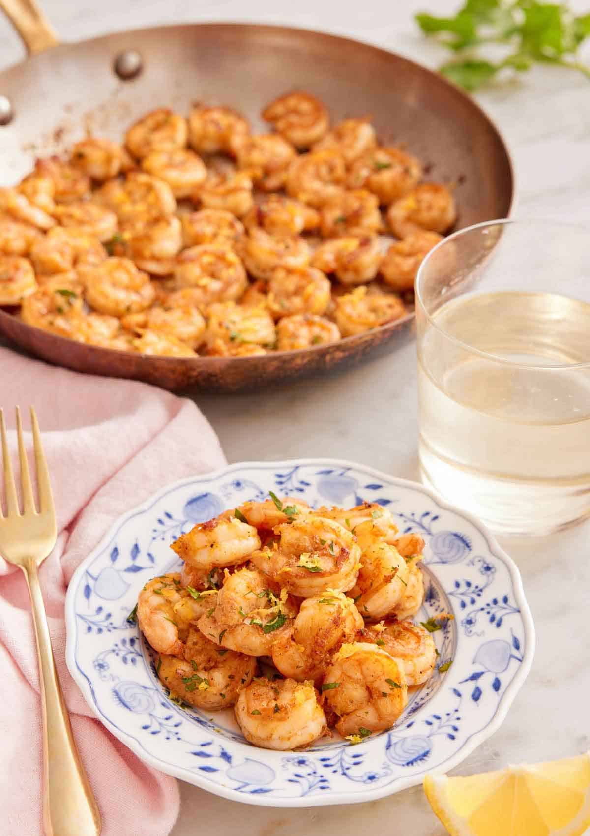 A plate of sautéed shrimp with a glass of wine and skillet of shrimp in the back.
