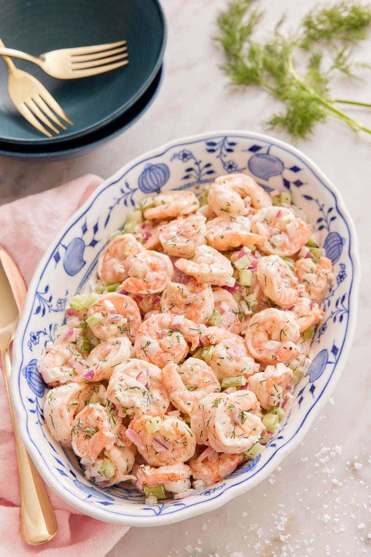 Overhead view of an oval platter of shrimp salad with some dill and stacked bowls and forks in the background.