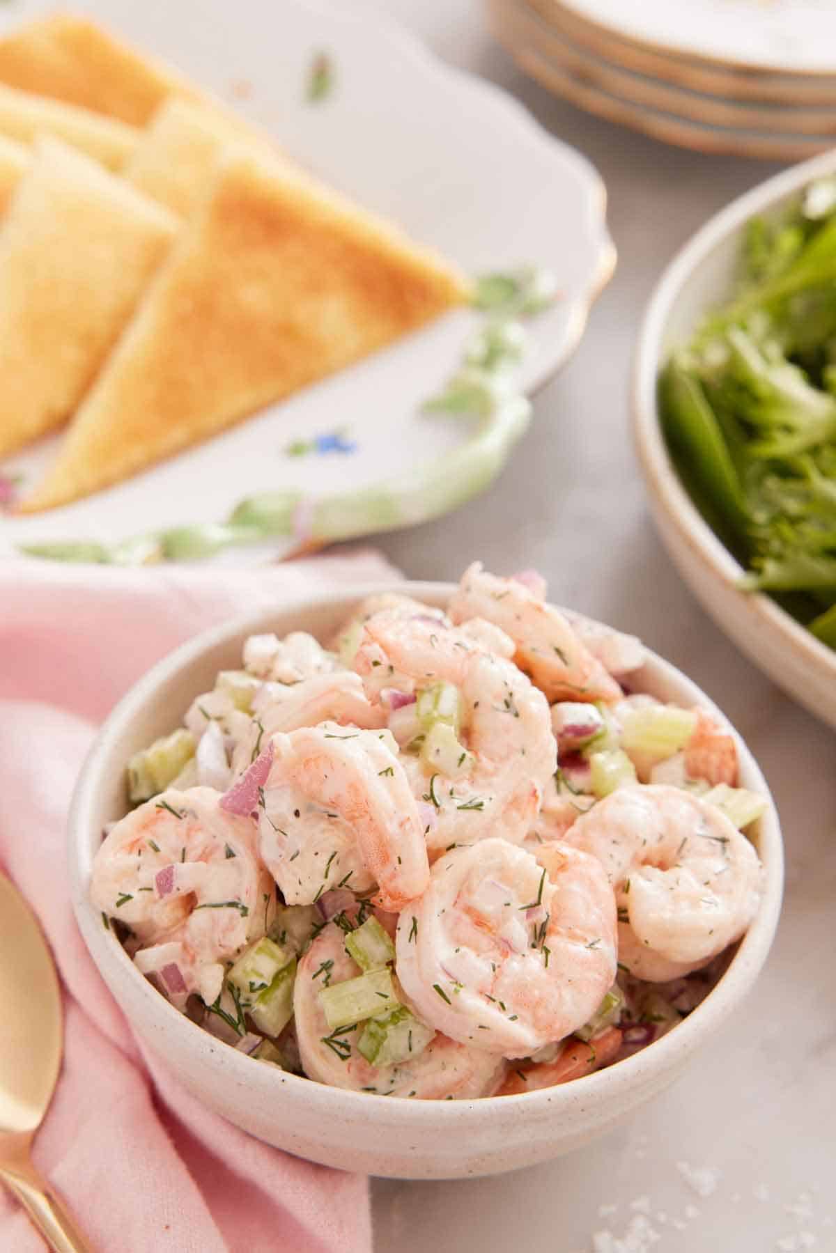 A bowl of shrimp salad with a plate of bread in the back.