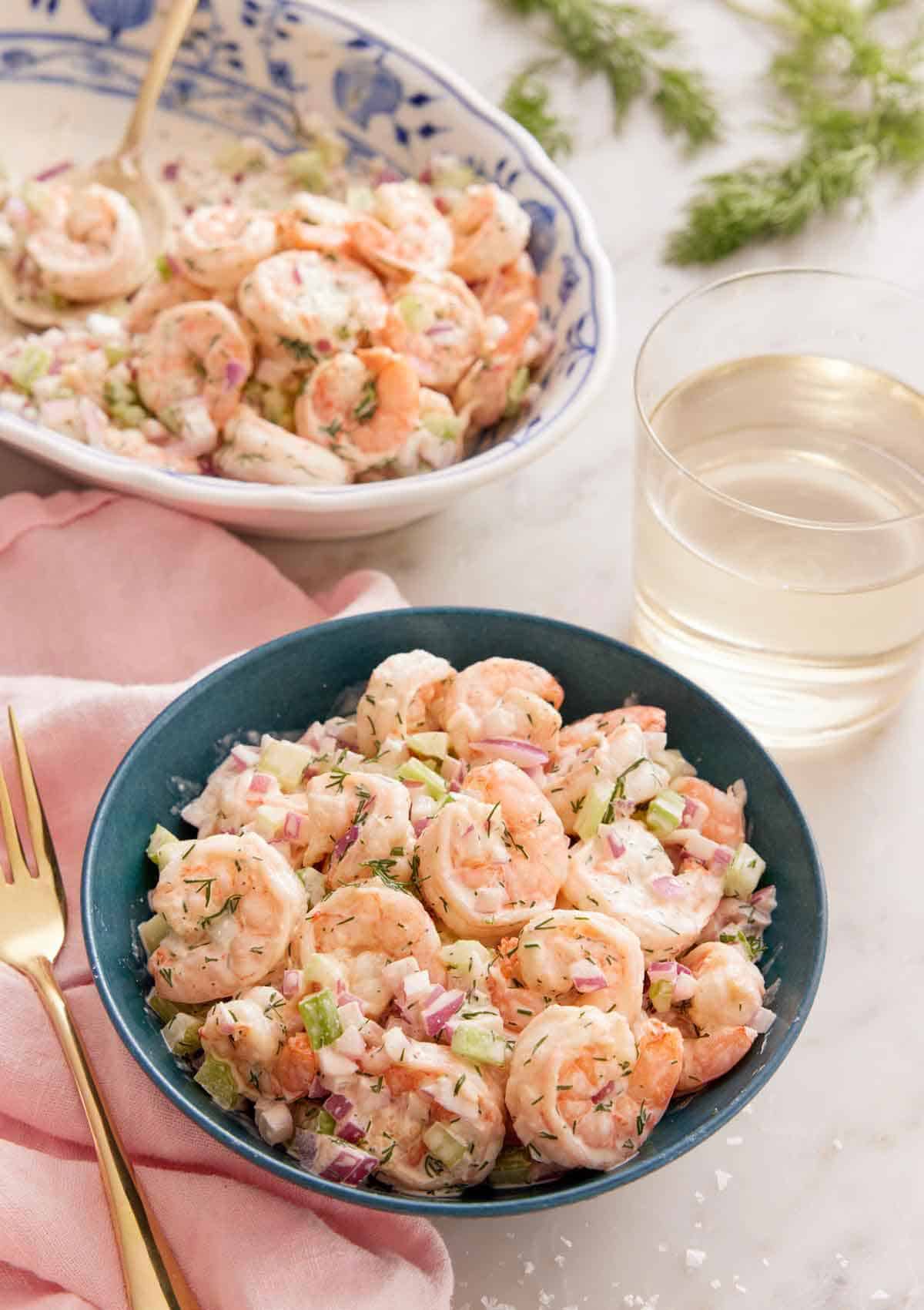 A bowl of shrimp salad with a glass of wine and platter of more shrimp in the background.