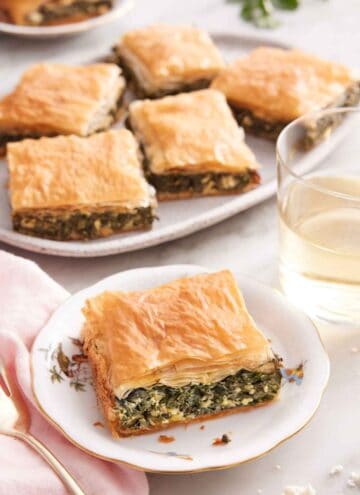 A plate with a square piece of spanakopita with a platter with more in the background.