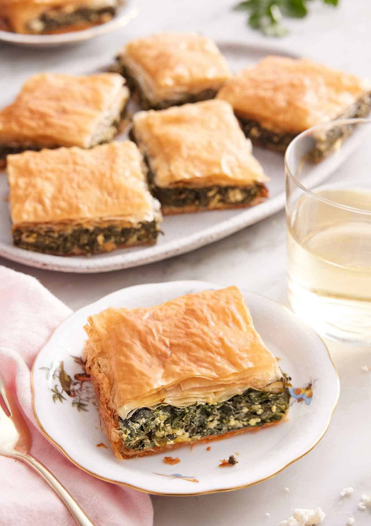 A plate with a square piece of spanakopita with a platter with more in the background.