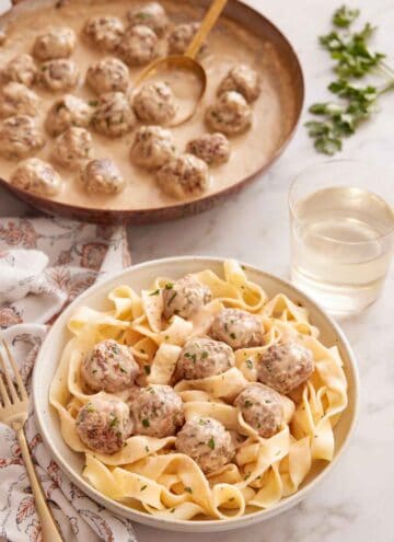 A bowl of eggs with Swedish meatballs on top with a skillet of more meatballs and a glass of wine behind it.