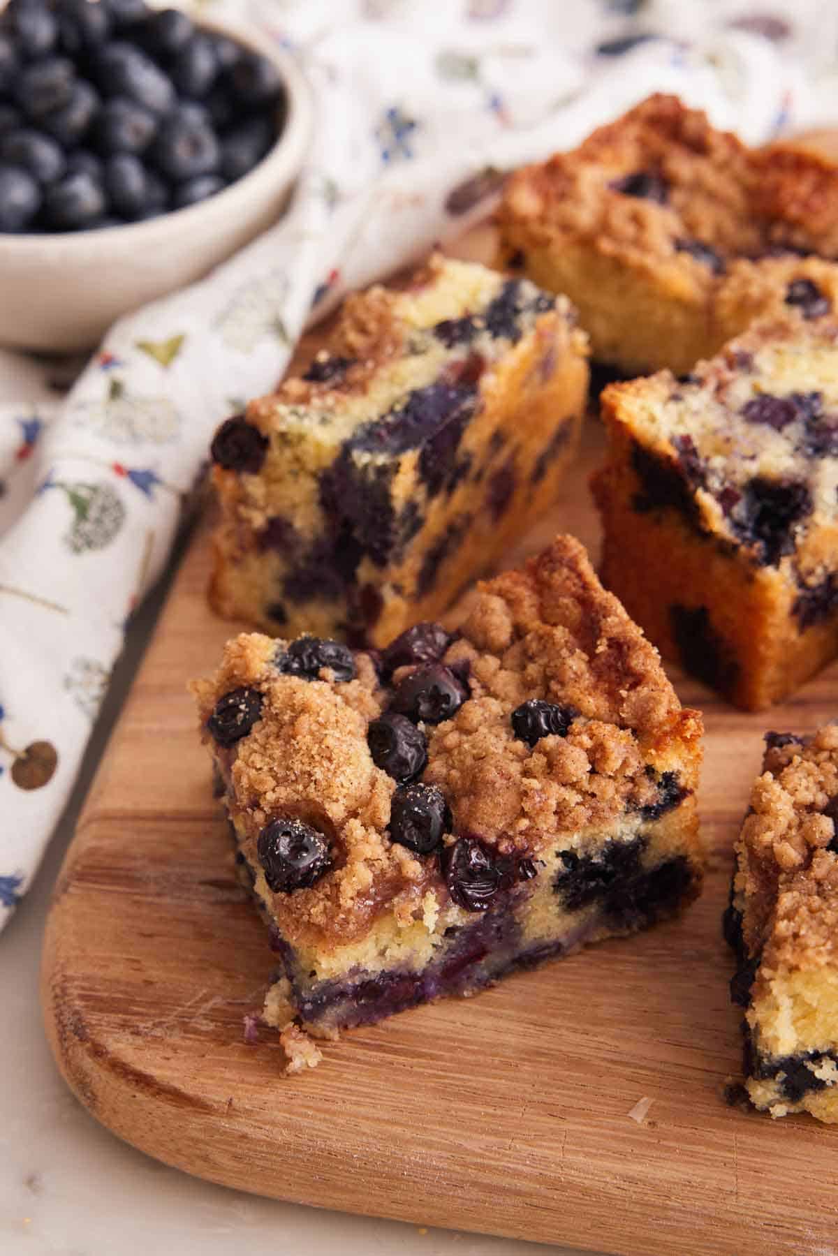 Multiple pieces of cut blueberry buckle on a cutting board with a bowl of blueberries in the background.